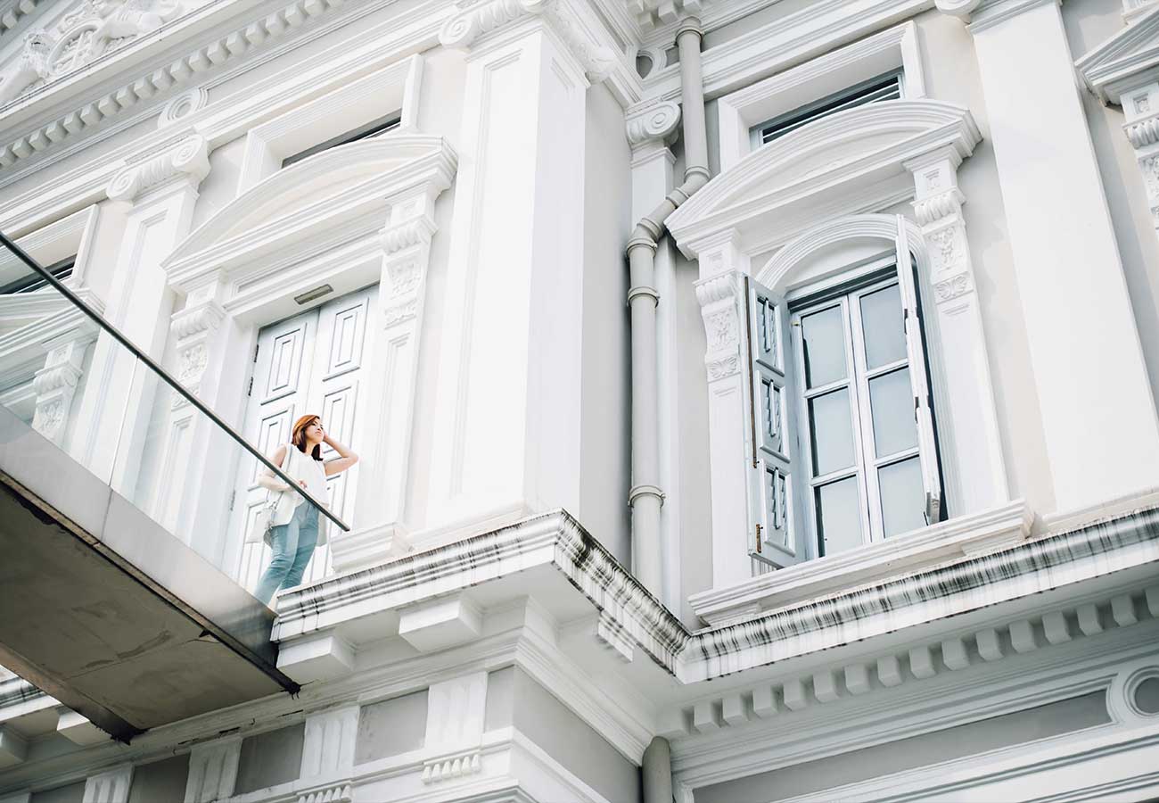 Woman stands on a glass balcony outside a white ornate building, looking up with hand to hair.