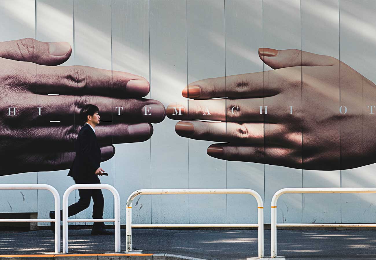 A businessman walks past a mural of two enormous hands reaching toward each other on a blue wall (handshake imagery).