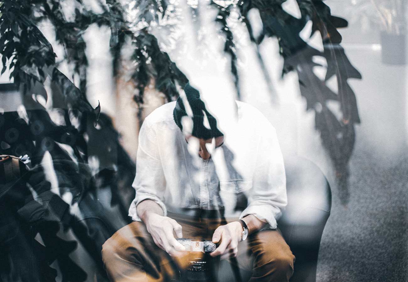 Person in a white shirt and brown pants sits, holding a camera, photographed through a glass window with plant reflections.