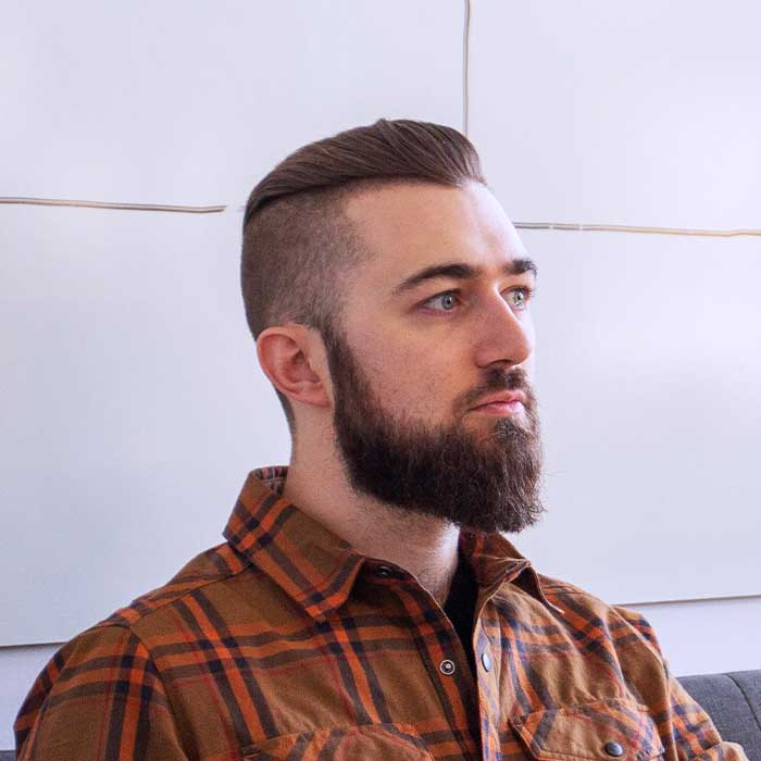 Portrait of a man with a full beard and slicked-back hair wearing a brown plaid shirt, looking to the right.