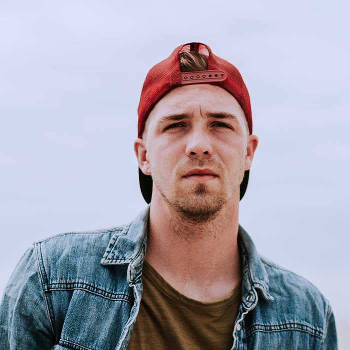 Man wearing a red backward cap and denim jacket, looking straight at the camera outdoors.