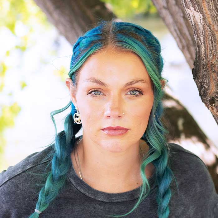Portrait of a woman with teal blue-dyed hair in two braids, posing outdoors beside a tree and looking at the camera with a neutral expression.