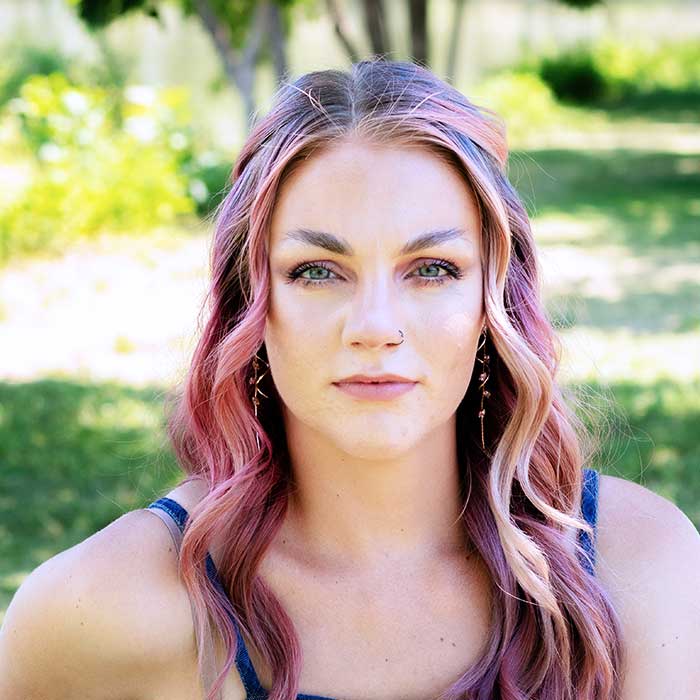 Portrait of a young woman with wavy pink‑purple hair outdoors, looking at the camera with a neutral expression; wearing a nose ring and earrings, in a sunlit park.