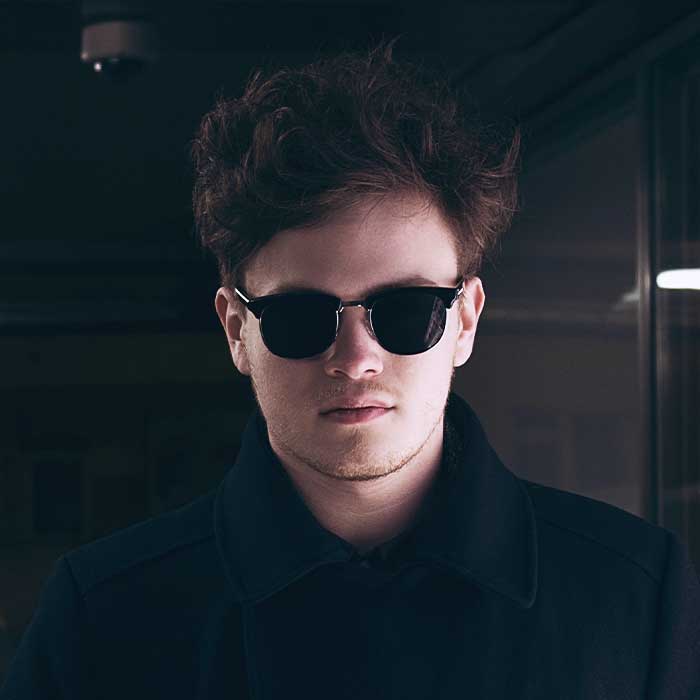 Portrait of a young man wearing dark sunglasses and a black coat in a moody, dimly lit setting.