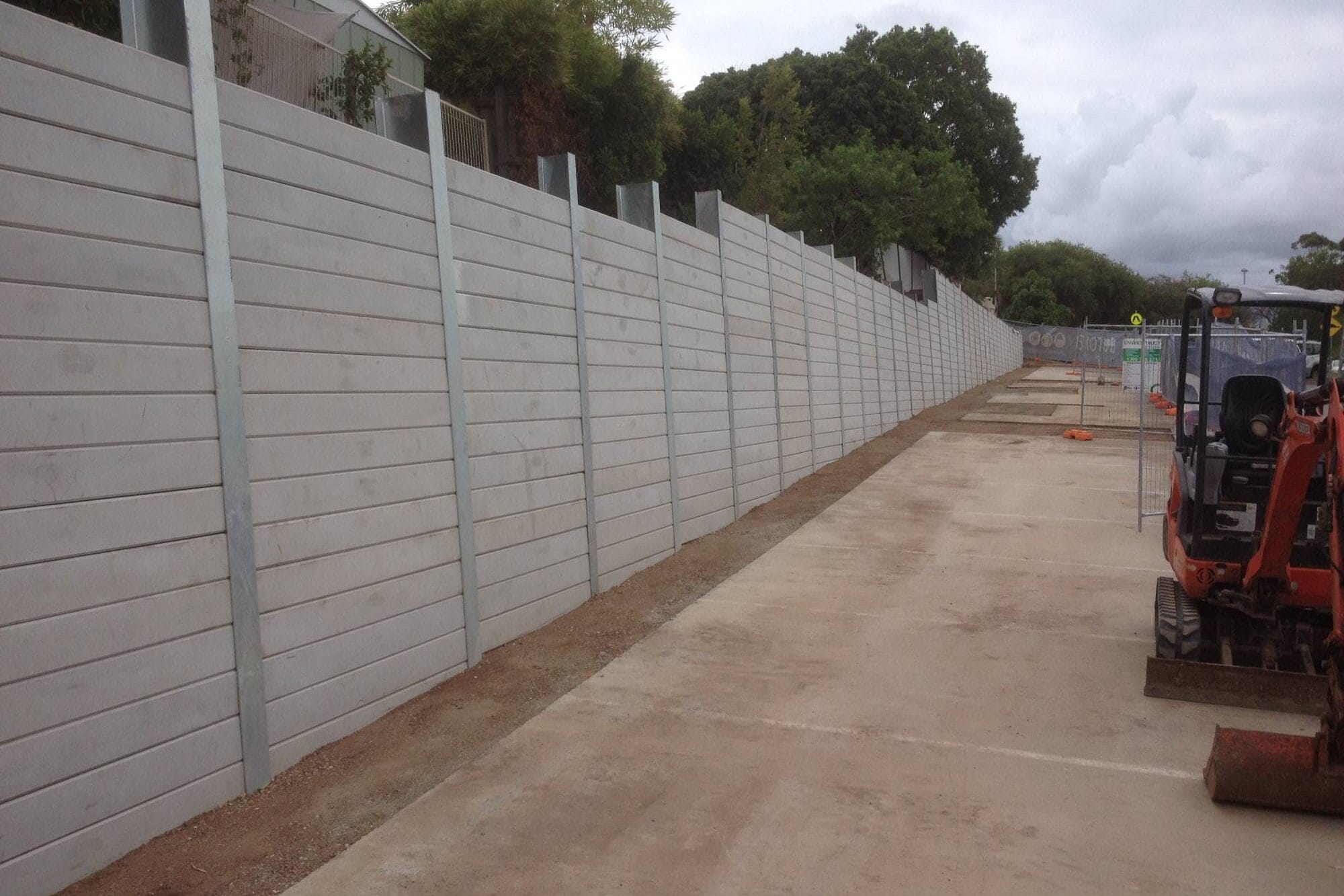 Civl Retaining Wall Geelong - Outwest Retaining Walls Long gray construction fence lining a sidewalk, with an orange excavator on the right and cloudy sky above
