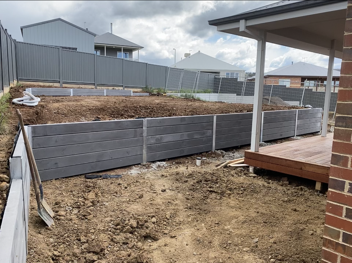 Backyard under construction with long gray raised garden beds along a fence and a wooden deck nearby, dirt soil visible.