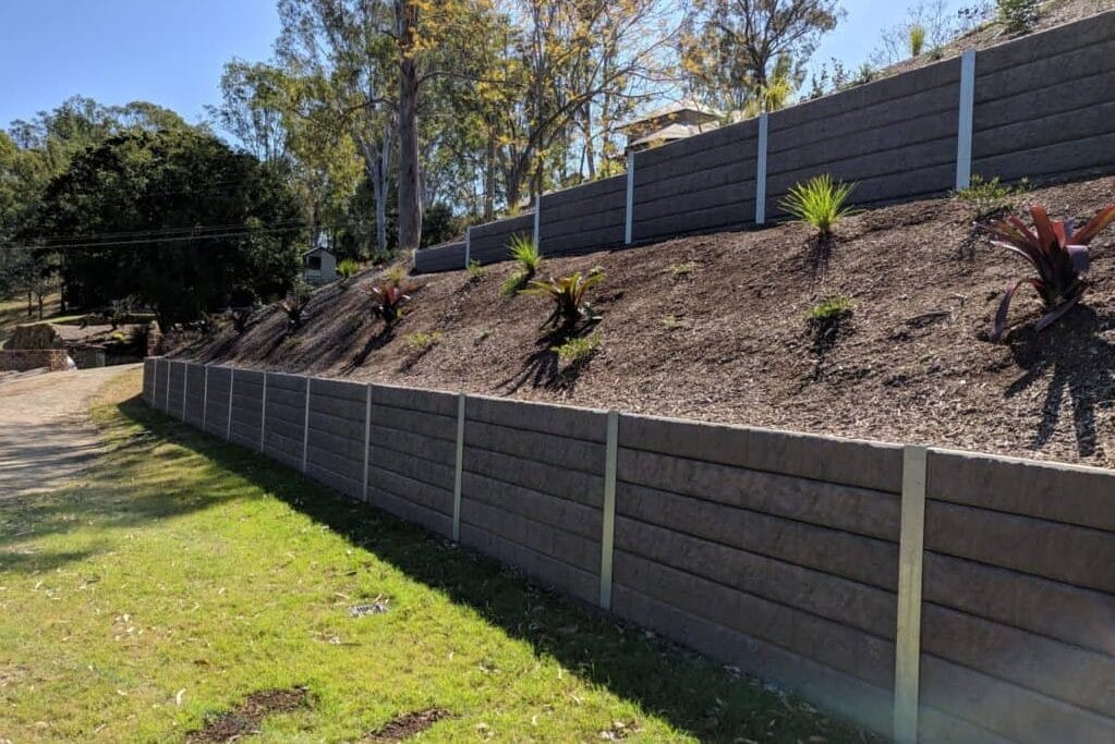 Retaining wall with Surcharge load - Outwest Retaining Walls Sloped garden bed behind stacked gray retaining walls with mulch and small plants on a sunny day.