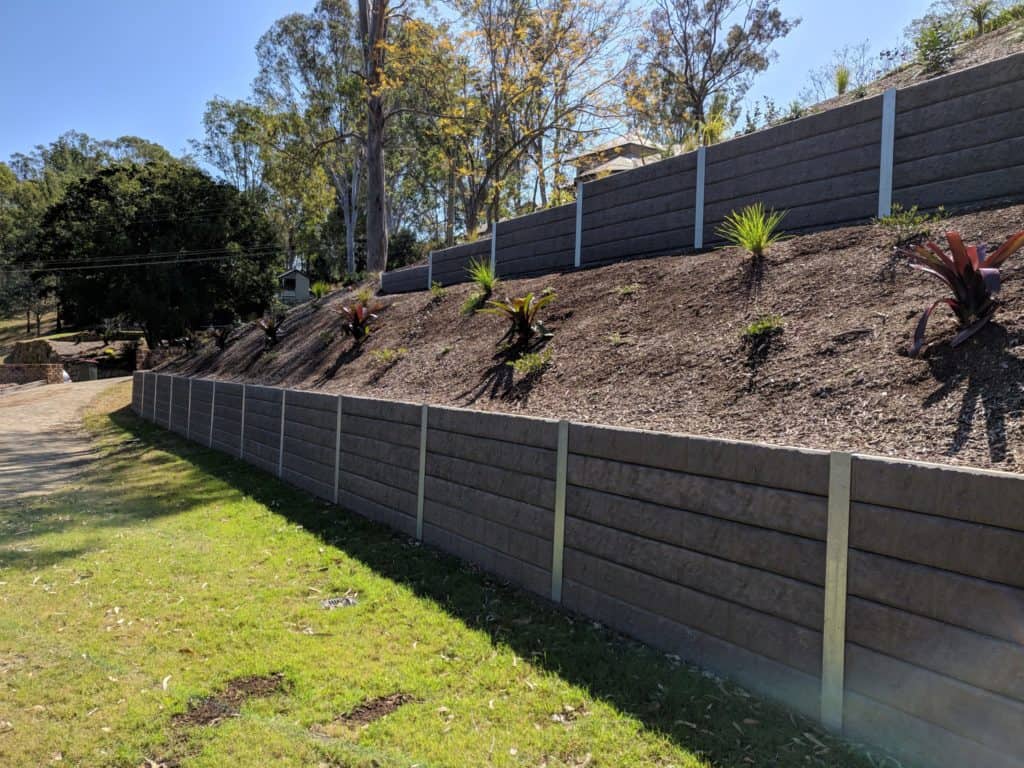 Sloped garden bed behind stacked gray retaining walls with mulch and small plants on a sunny day.
